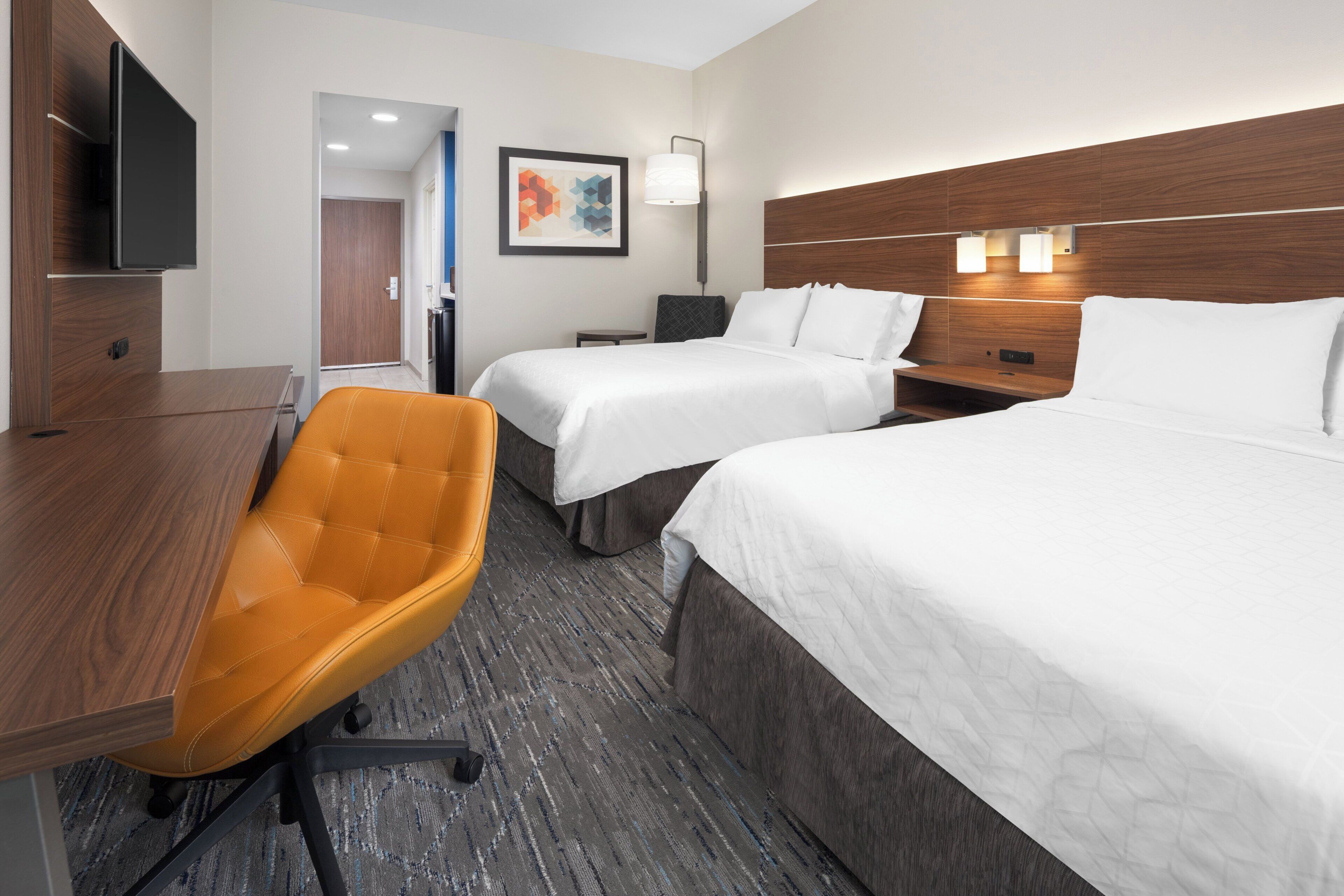 A hotel room with two beds, a desk, a chair, a TV, and a wall-mounted lamp.