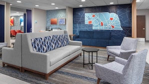 Un hall d’entrée avec un canapé gris, un divan bleu et un fauteuil gris. On y trouve une petite table et un mur décoré d’une carte.