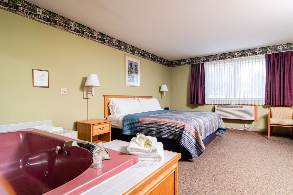 A hotel room with a large bathtub, a bed with a blue and white striped blanket, a wooden nightstand, a chair, and a window with purple curtains.