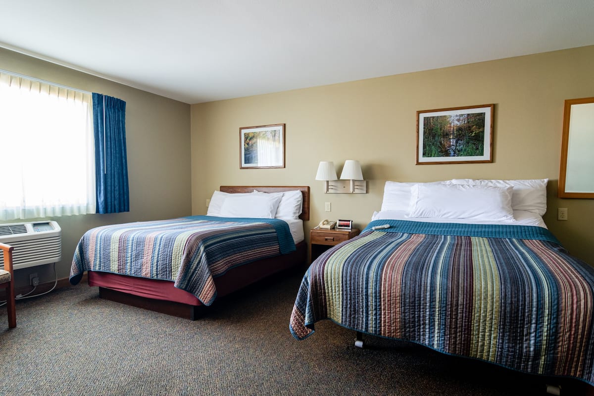 A hotel room with two beds, a window with curtains, an air conditioning unit, a nightstand with lamps, and two framed pictures on the wall.