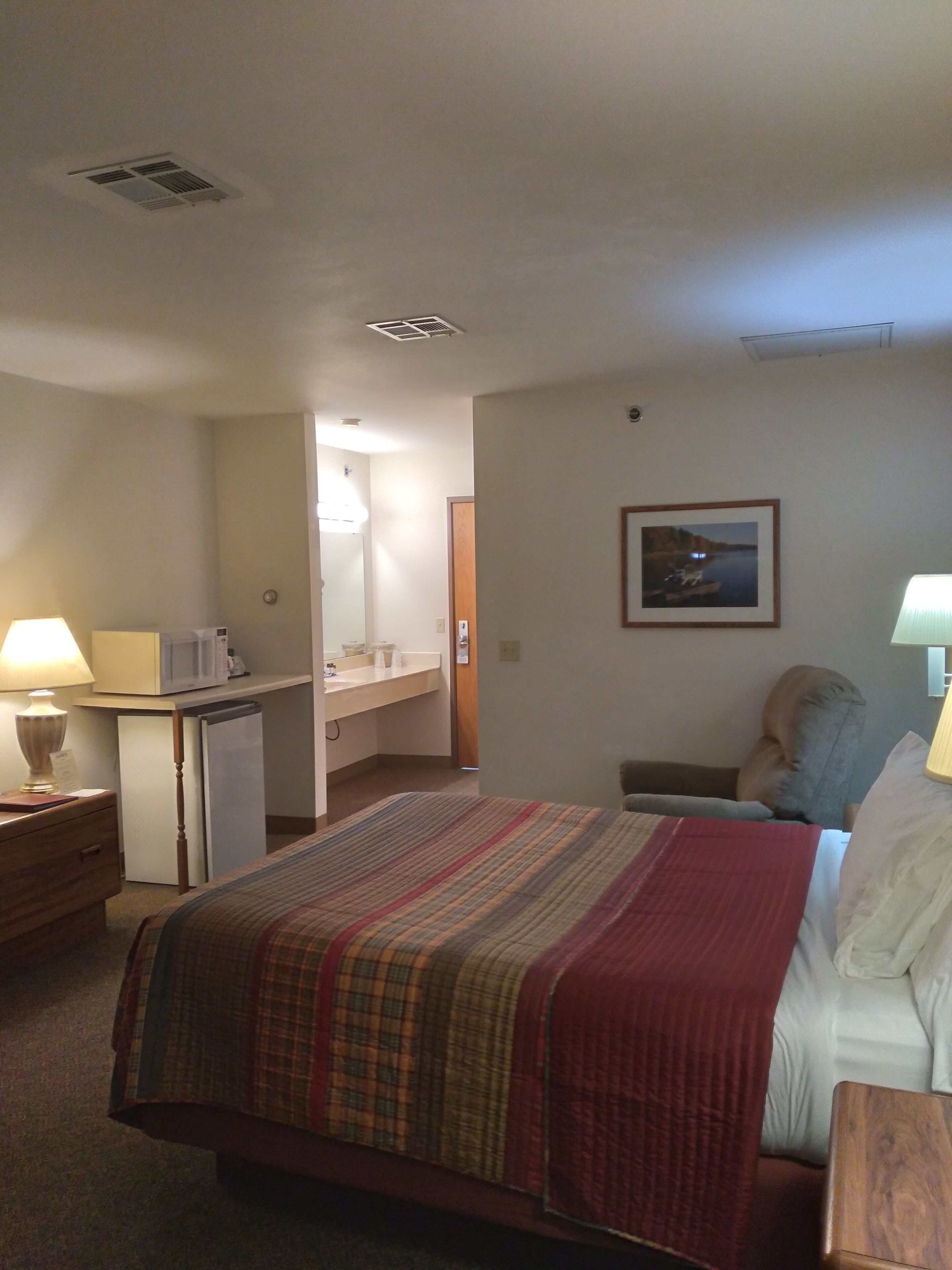 Habitación de hotel con cama, escritorio, microondas, refrigerador, mesita de noche, lámpara, silla y un cuadro en la pared.