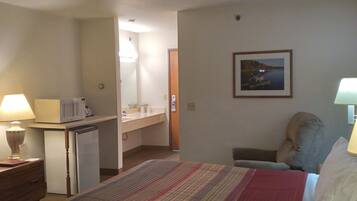 Habitación de hotel con cama, escritorio, microondas, refrigerador, mesita de noche, lámpara, silla y un cuadro en la pared.