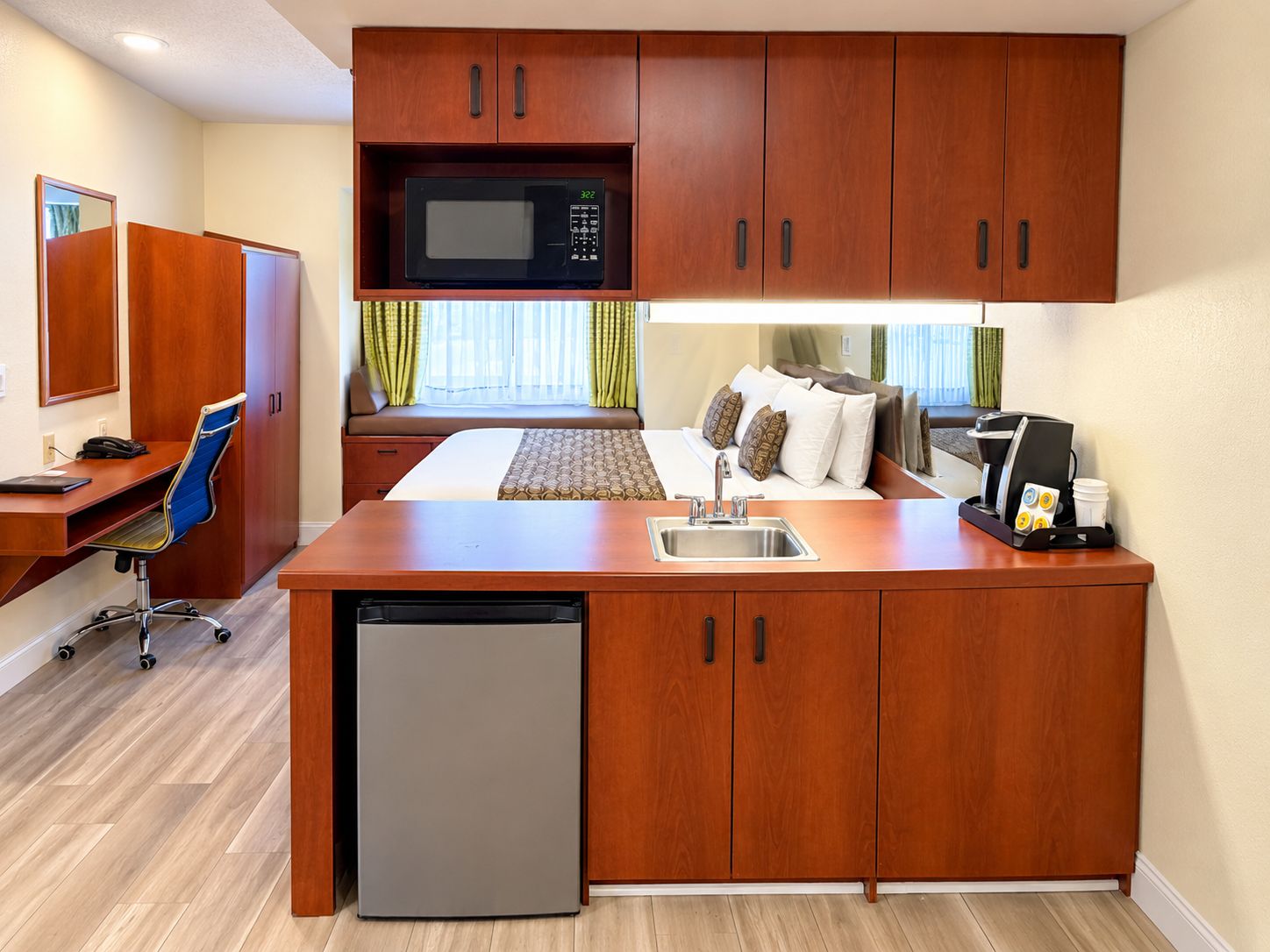 Habitación de hotel compacta con kitchenette, cama con vistas, escritorio con silla y un refrigerador pequeño.