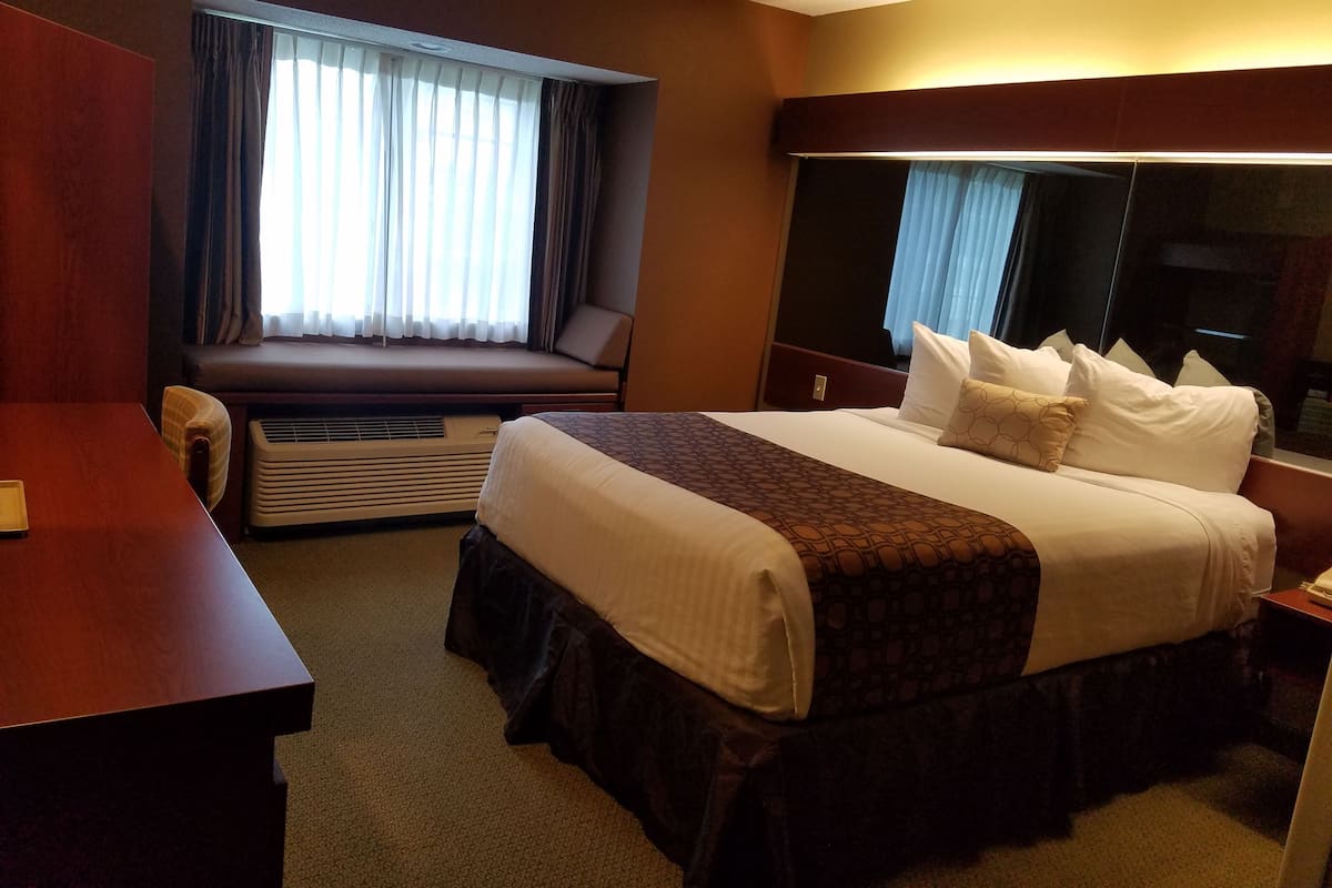 A hotel room with a large bed, a desk, a window with curtains, an air conditioning unit, and a mirror.