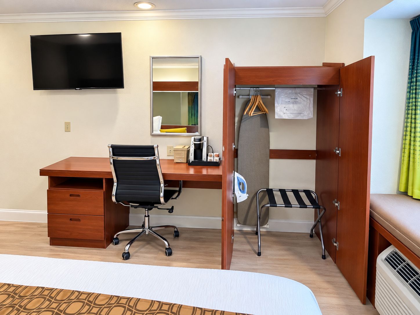 Une chambre d’hôtel avec un bureau en bois, un fauteuil de bureau noir, un téléviseur à écran plat fixé au mur, un miroir, une armoire avec une tringle à vêtements et un lit recouvert d’une couvre-lit à motifs.