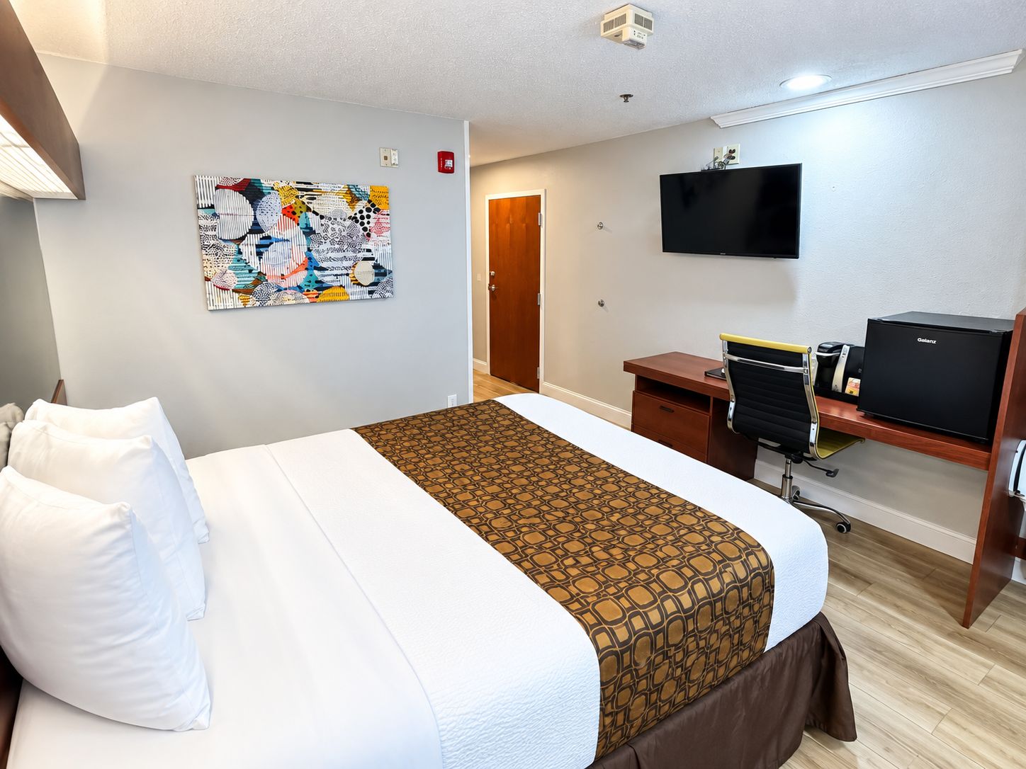 Habitación de hotel con cama, escritorio con computadora, televisor de pantalla plana y una obra de arte colgada en la pared.