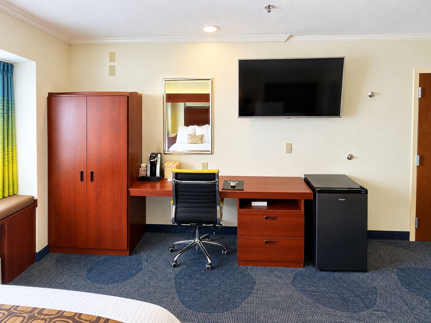 A hotel room with a bed, a desk with a computer, a chair, a wardrobe, a television, and a minibar.