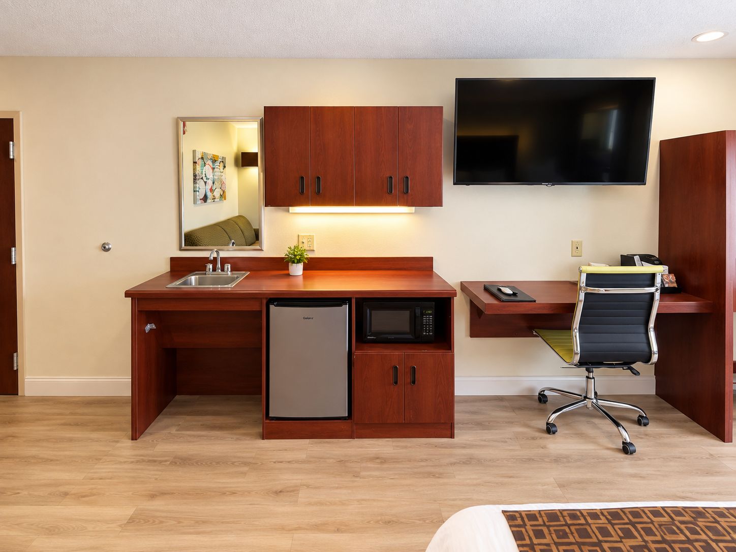 A compact hotel room with a kitchenette, a flat-screen TV, a desk, and a chair.