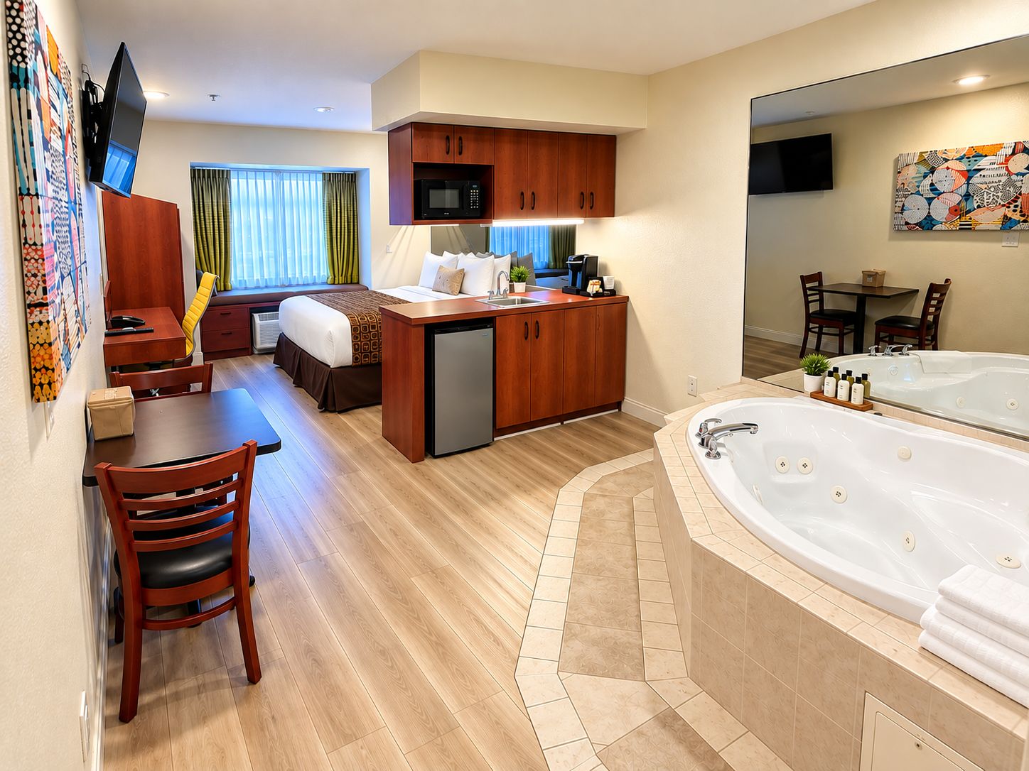 Een hotelkamer met een kitchenette, een eethoek, een bed, een badkamer met een groot bad en een aan de muur bevestigde televisie.