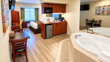 A hotel room with a kitchenette, a dining area, a bed, a bathroom with a large bathtub, and a TV mounted on the wall.