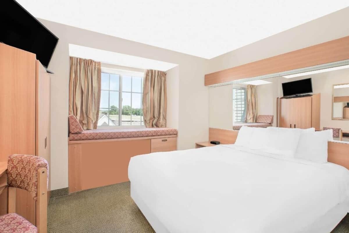 A hotel room with a large bed, a desk with a chair, a window with curtains, a television, and a mirror.