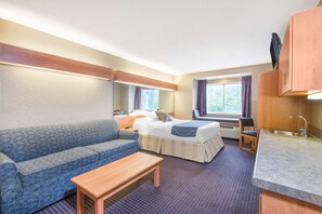 Een kleine hotelkamer met een bed, een slaapbank, een tafeltje, een kitchenette en een raam met gordijnen.