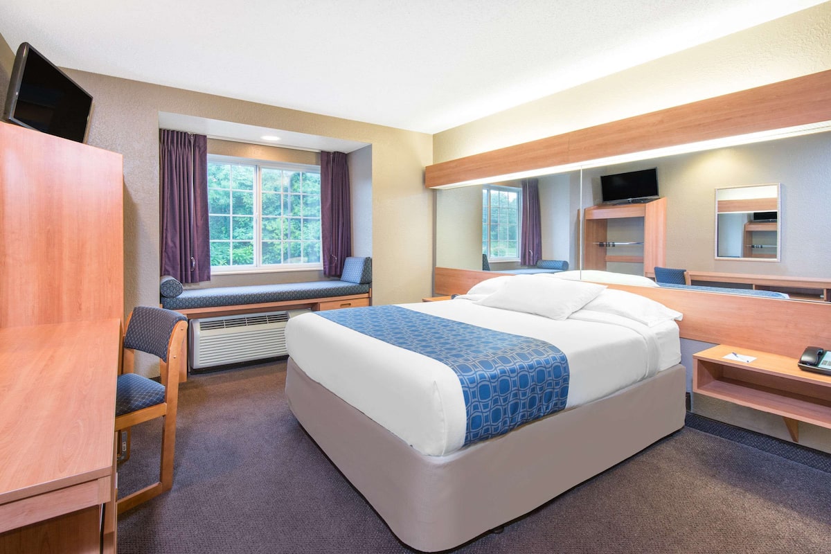 Ein Hotelzimmer mit Bett, Schreibtisch, Stuhl, einem Fenster mit Vorhängen, einem Fernseher und einem Spiegel.