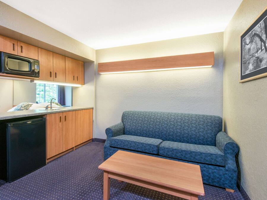 A compact kitchenette with a microwave, sink, and dishwasher, a blue patterned sofa, and a wooden table.