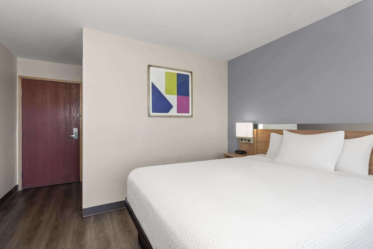 Habitación de hotel con una cama grande, una puerta de madera, una mesita de noche con lámpara y una obra de arte abstracta colgada en la pared.