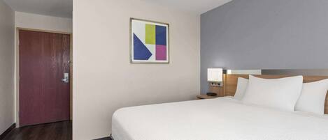 A hotel room with a large bed, a wooden door, a nightstand with a lamp, and a wall-mounted abstract art piece.