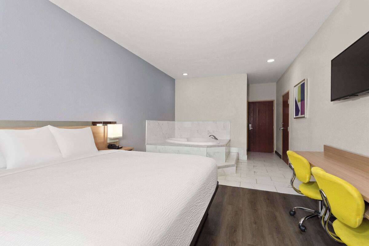 Habitación de hotel con una cama grande, baño con bañera, escritorio con sillas amarillas y televisión de pantalla plana montada en la pared.