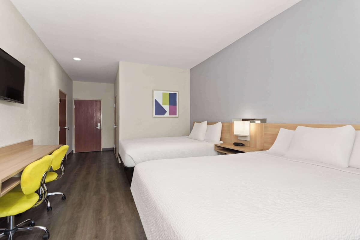 Habitación de hotel con dos camas, un escritorio con sillas, un televisor y un cuadro en la pared.