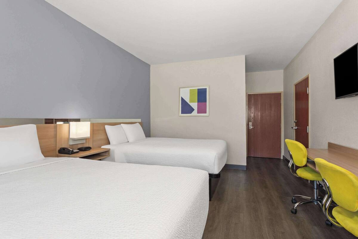 Habitación de hotel con dos camas, un escritorio con silla, televisor montado en la pared y una puerta que da a otra habitación.