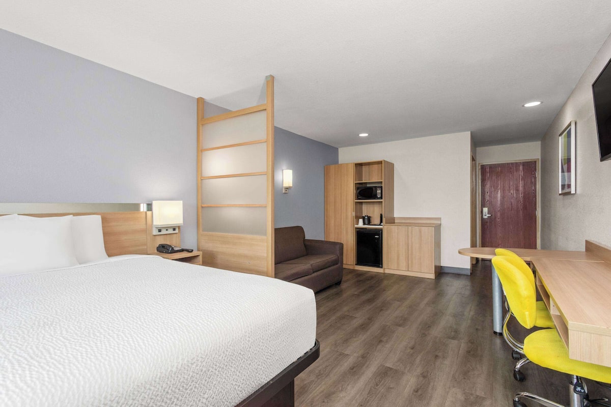 Habitación de hotel con cama, escritorio, silla, microondas, pequeña cocina americana y puerta corredera.