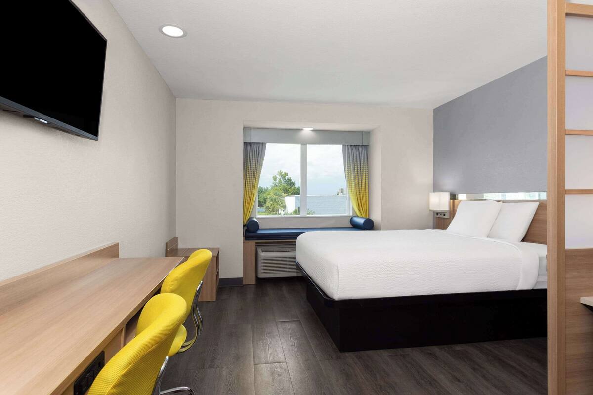 Habitación de hotel con cama, escritorio, silla, televisor y ventana con cortinas.