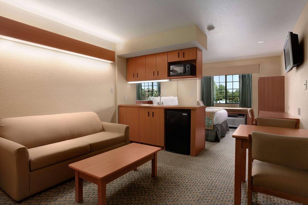 Habitación de hotel compacta con sofá, una mesita, kitchenette con microondas y televisor fijado a la pared.