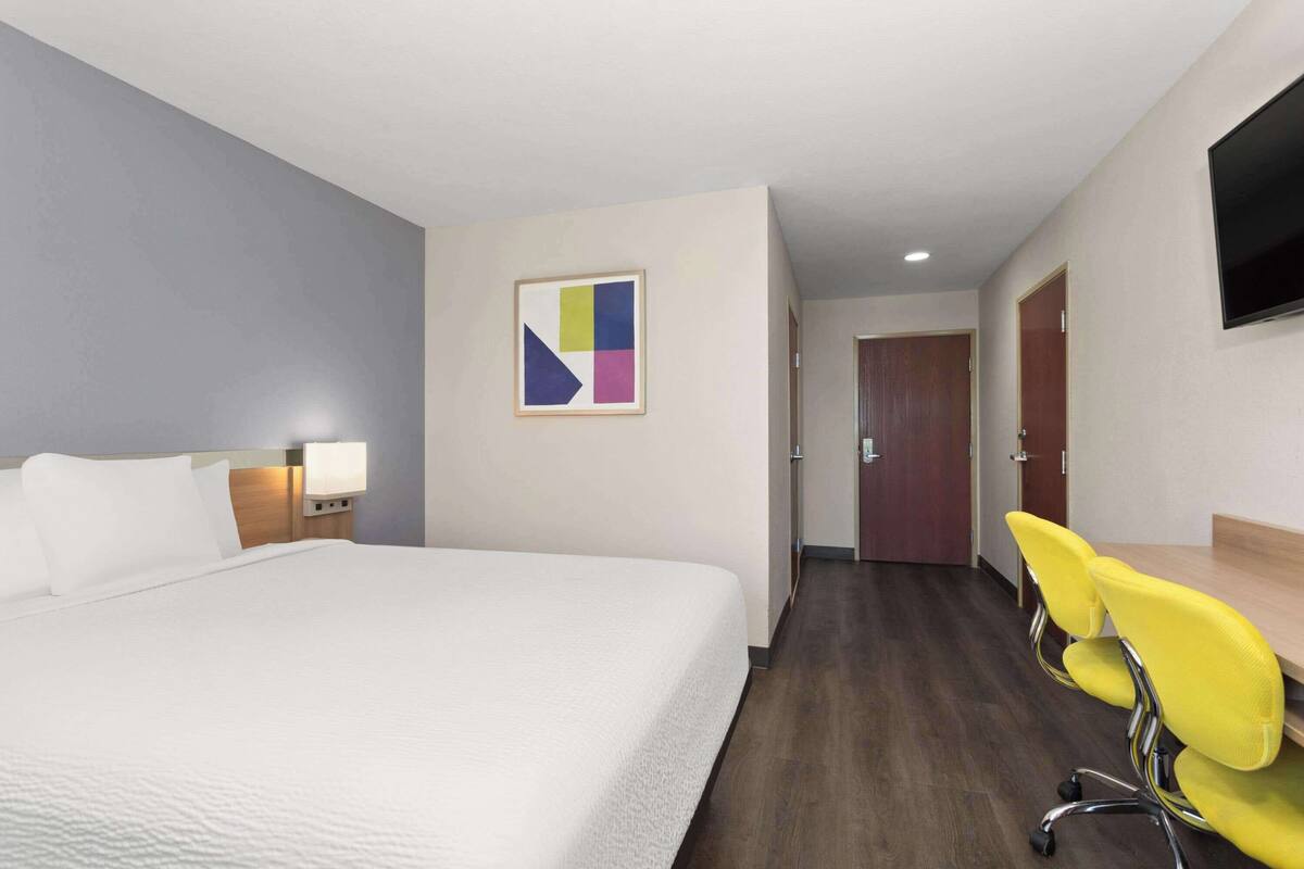 Habitación de hotel con una cama grande, un escritorio con dos sillas amarillas, un televisor de pantalla plana en la pared y un cuadro abstracto moderno.