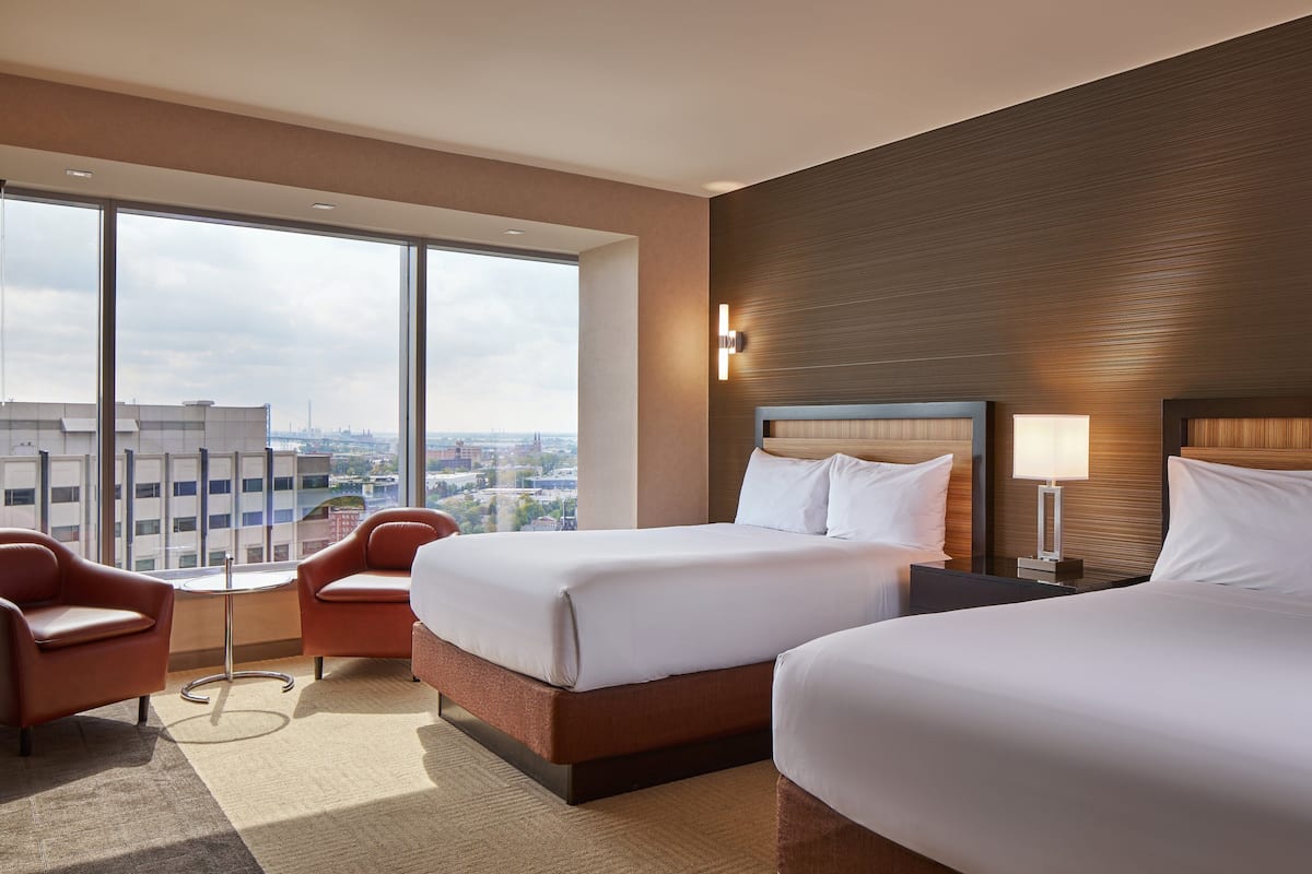A hotel room with two beds, a large window with a city view, a small table, and a chair.