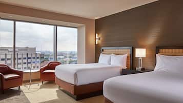 A hotel room with two beds, a large window with a city view, a small table, and a chair.