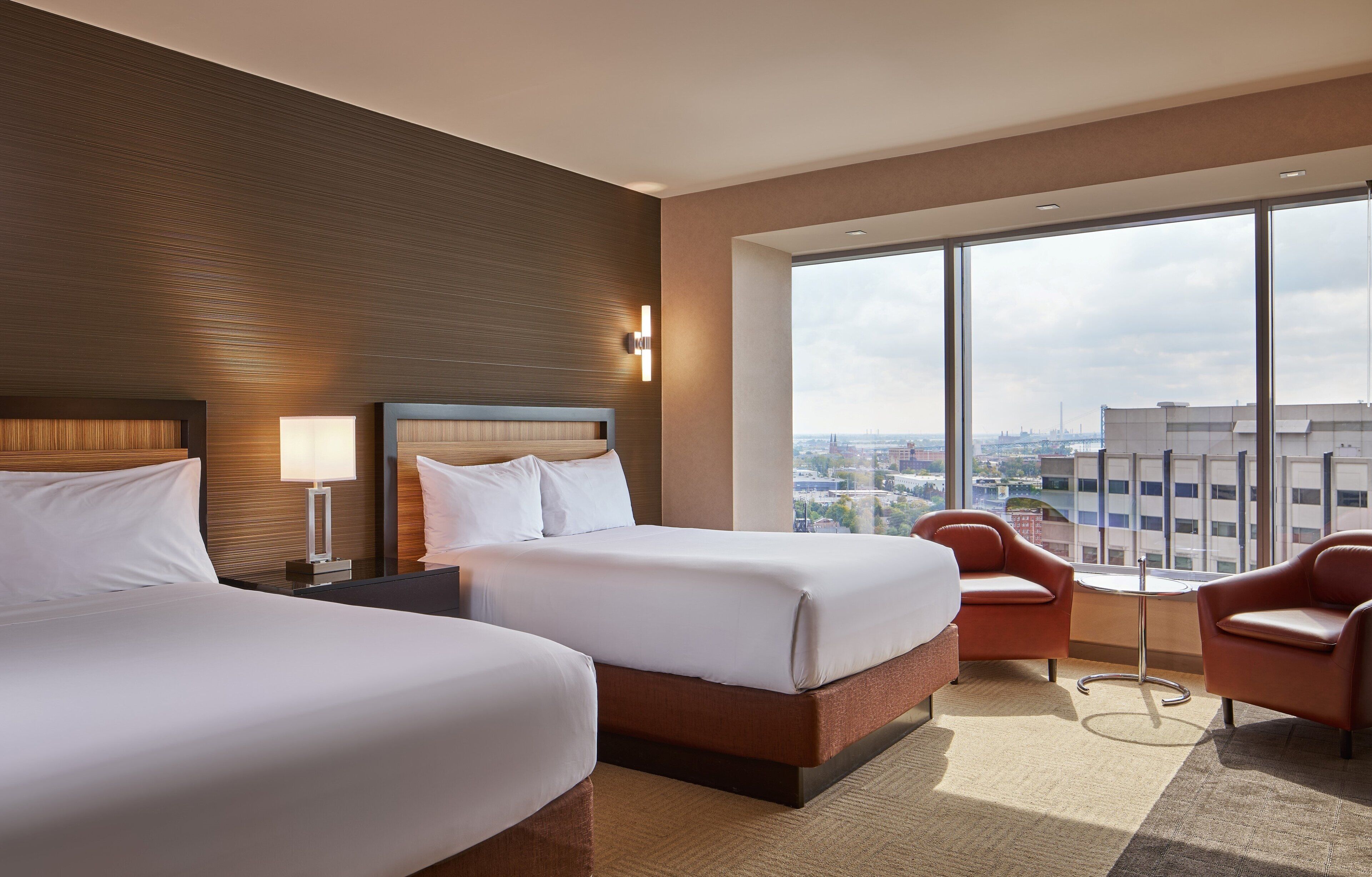 A hotel room with two beds, a large window with a city view, and a small table with two chairs.