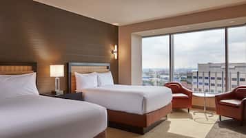A hotel room with two beds, a large window with a city view, and a small table with two chairs.