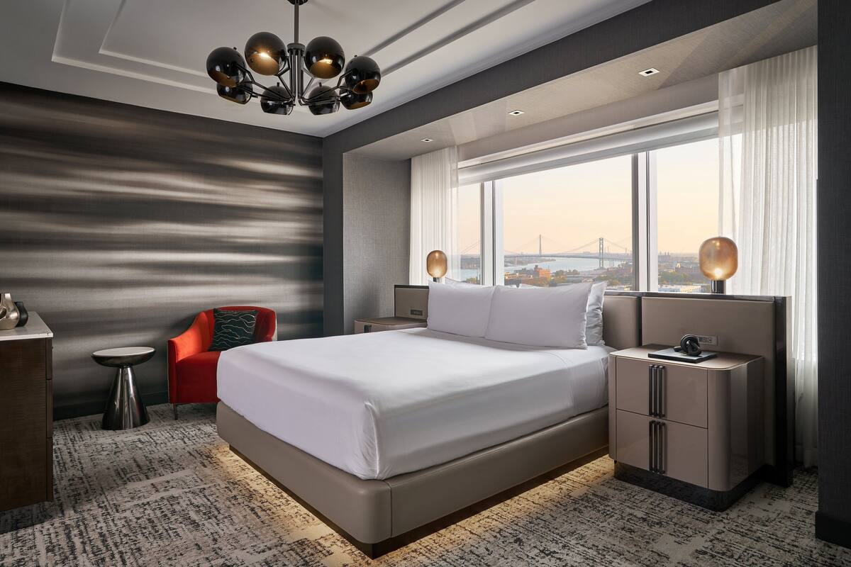 A modern hotel room with a large bed, a red chair, a small side table, and a nightstand with a lamp.