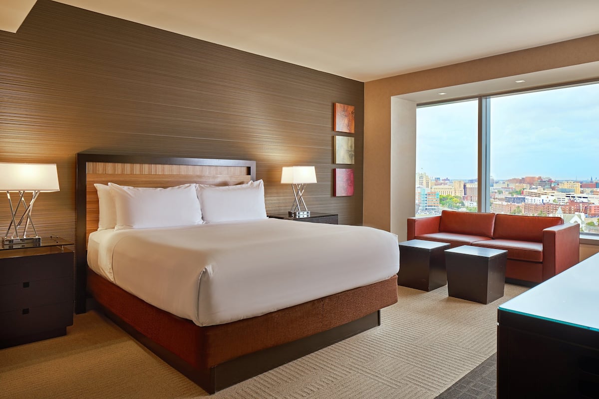 A hotel room with a large bed, a red sofa, a desk, and a view of the city through large windows.