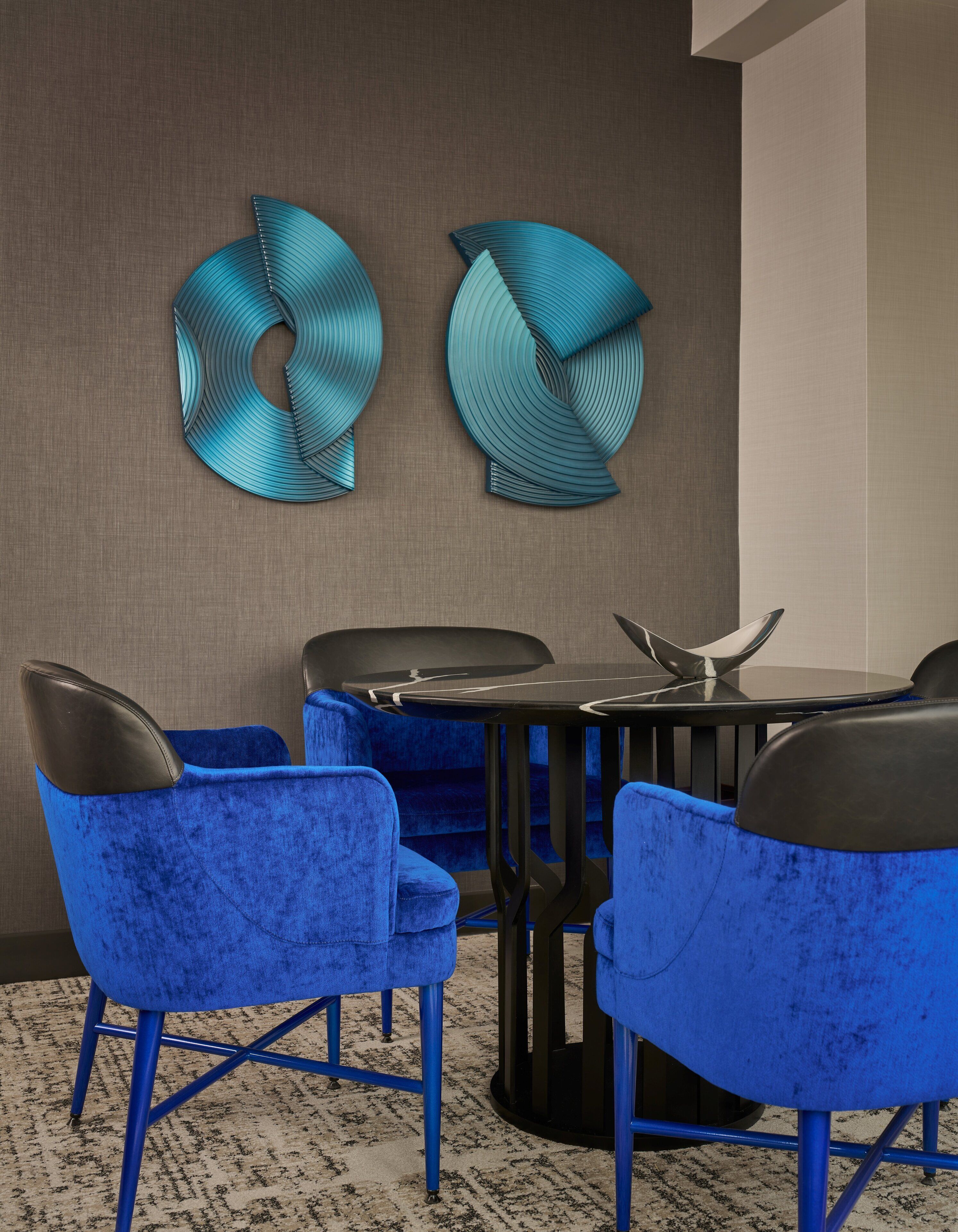 A modern room with blue velvet chairs, a black table, and two abstract wall art pieces.