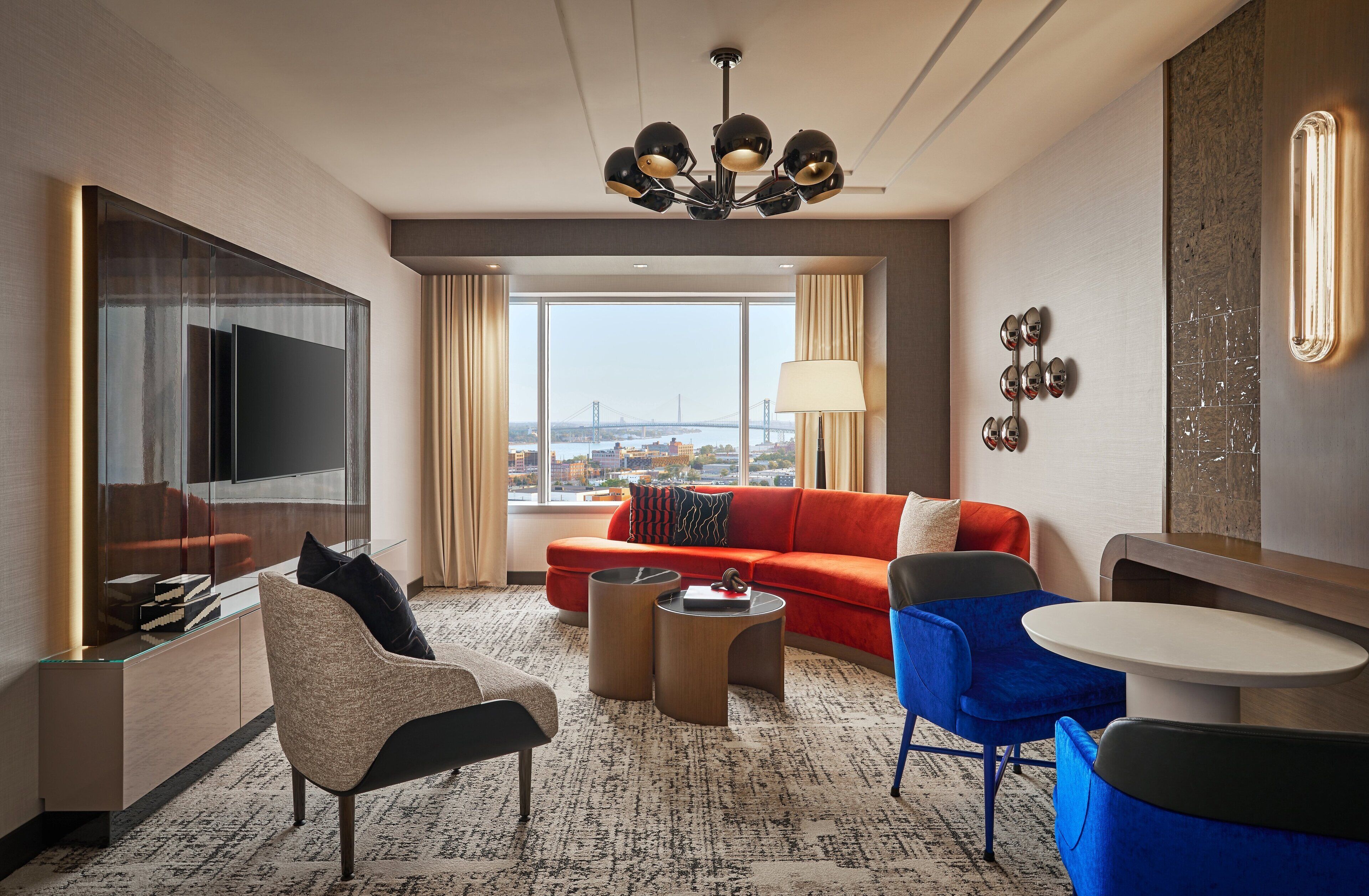 A modern hotel room with a large TV, a red sofa, a blue armchair, a small round table, and a view of a cityscape through large windows.
