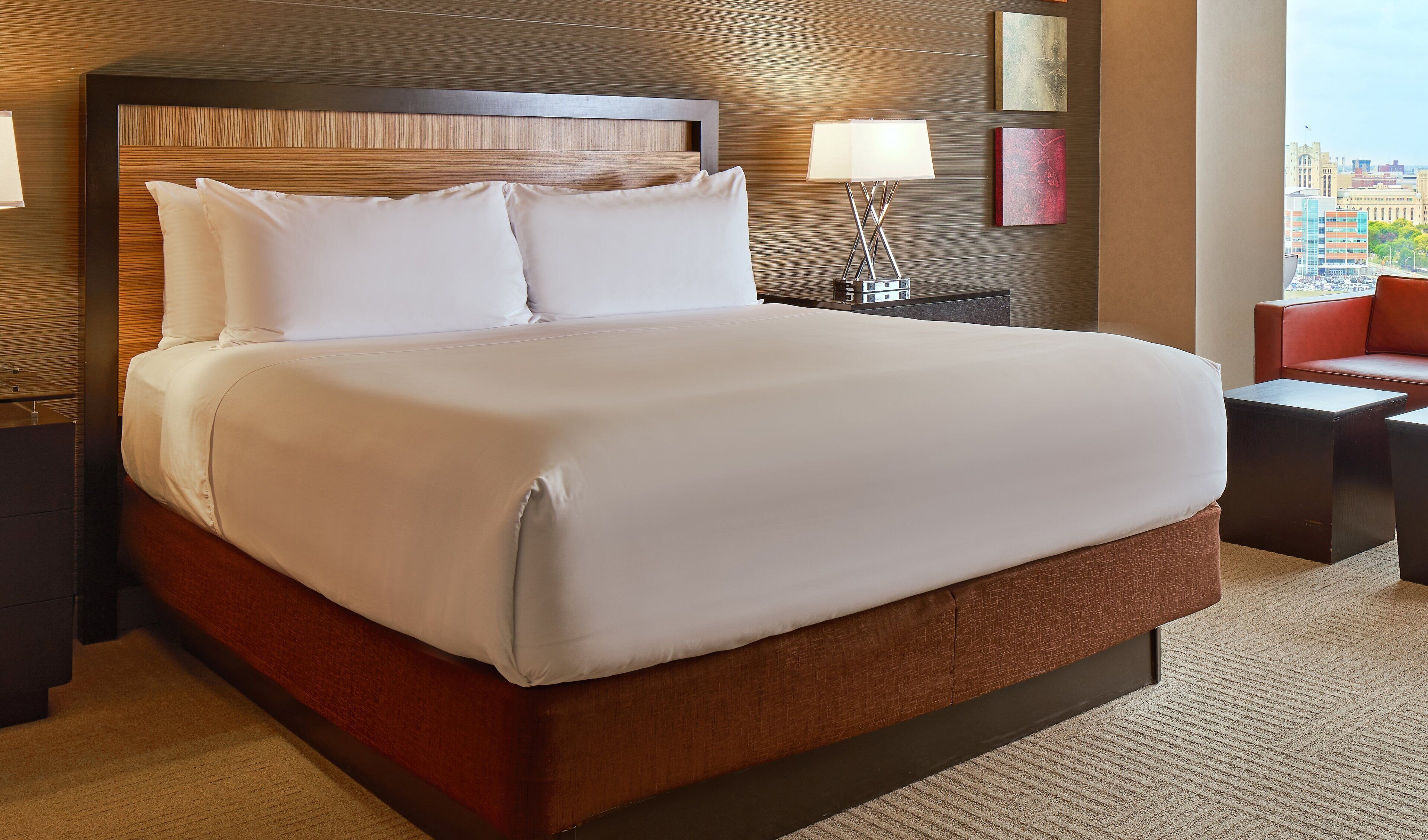 Een hotelkamer met een groot bed, een nachtkastje met een lamp, een rode stoel en uitzicht op de stad.