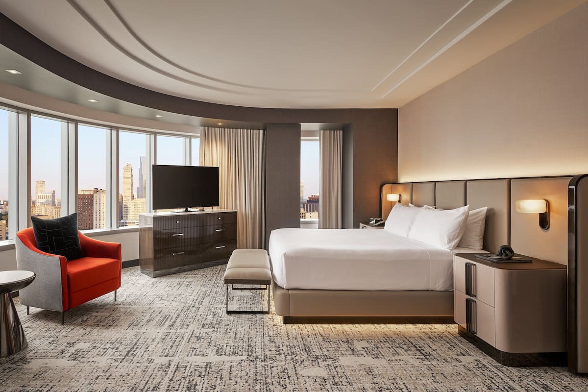 A modern hotel room with a large bed, a TV, a desk, and a view of the city skyline.