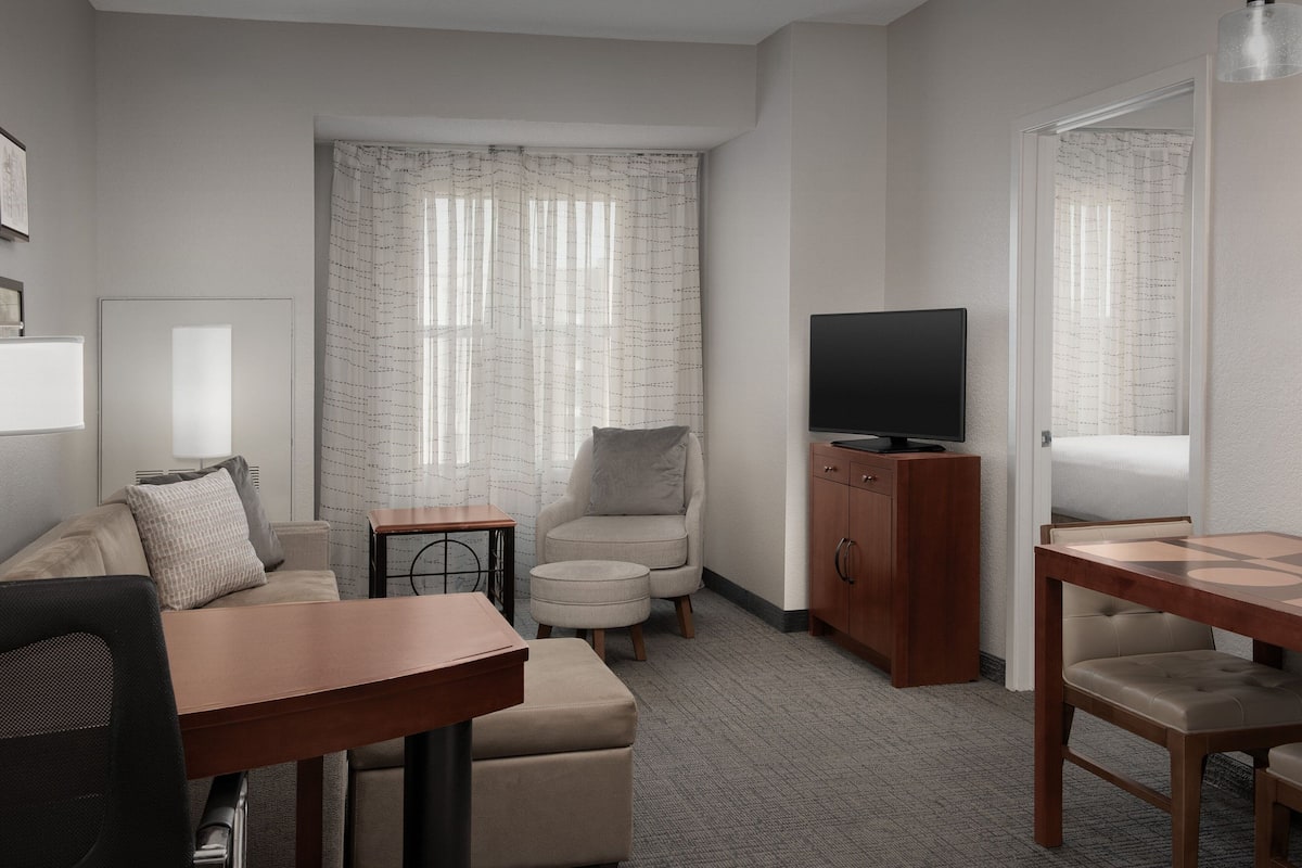 A modern hotel room with a sofa, armchair, small table, TV, and a wooden cabinet.