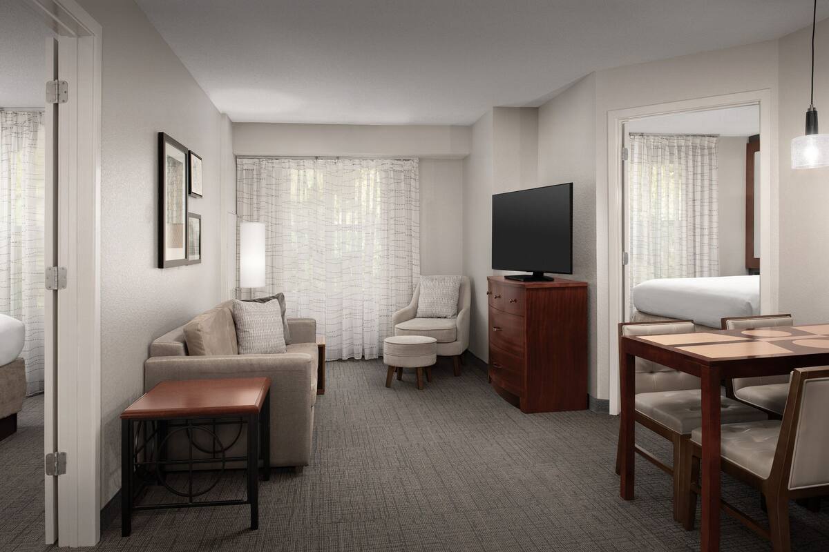 A modern hotel room with a sofa, armchair, ottoman, wooden side table, TV, and a dining area with a table and chairs.