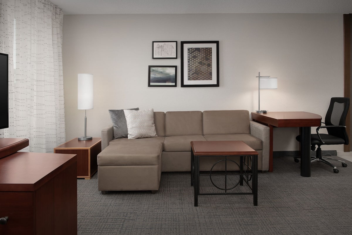 A modern hotel room with a sectional sofa, a wooden desk, a standing lamp, and framed artwork on the wall.