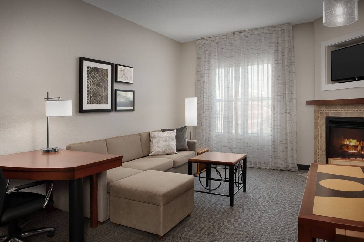 A modern hotel room with a sofa, a small table, a desk, a chair, a TV, and a fireplace.
