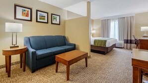 A hotel room with a blue sofa, a wooden coffee table, a bed with a green blanket, a lamp, and two framed pictures on the wall. - Comfort Inn Plover-Stevens Point (Plover)