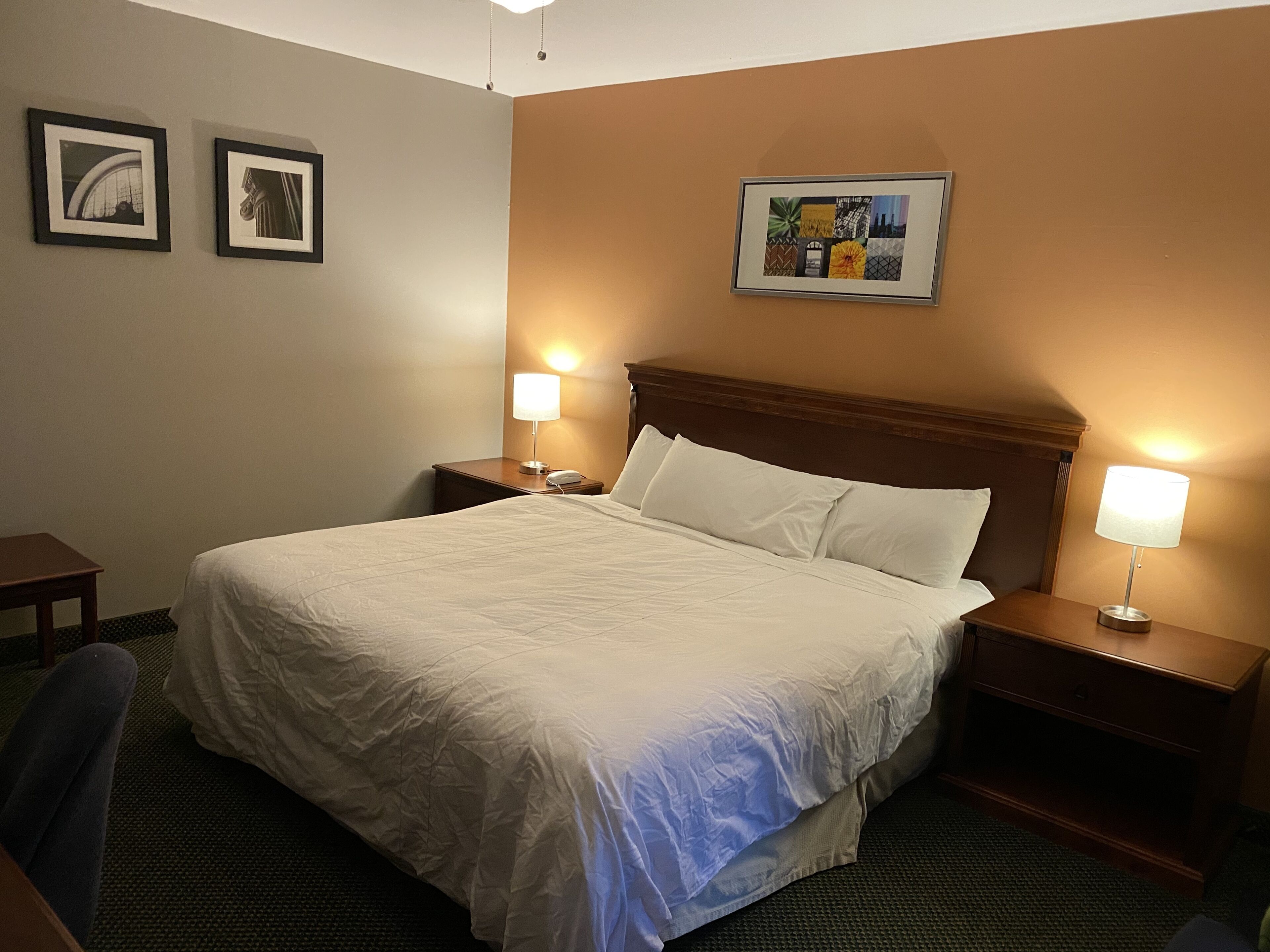 Hotellihuone, jossa on suuri sänky, kaksi yöpöytää, yölamppu, tuoli ja seinä, jossa on kolme kehystettyä kuvaa.