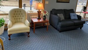Una sala de estar con un sillón color crema, un sofá oscuro, una pequeña mesa de madera con una lámpara y flores, y ventanas grandes con vistas a una calle y edificios.