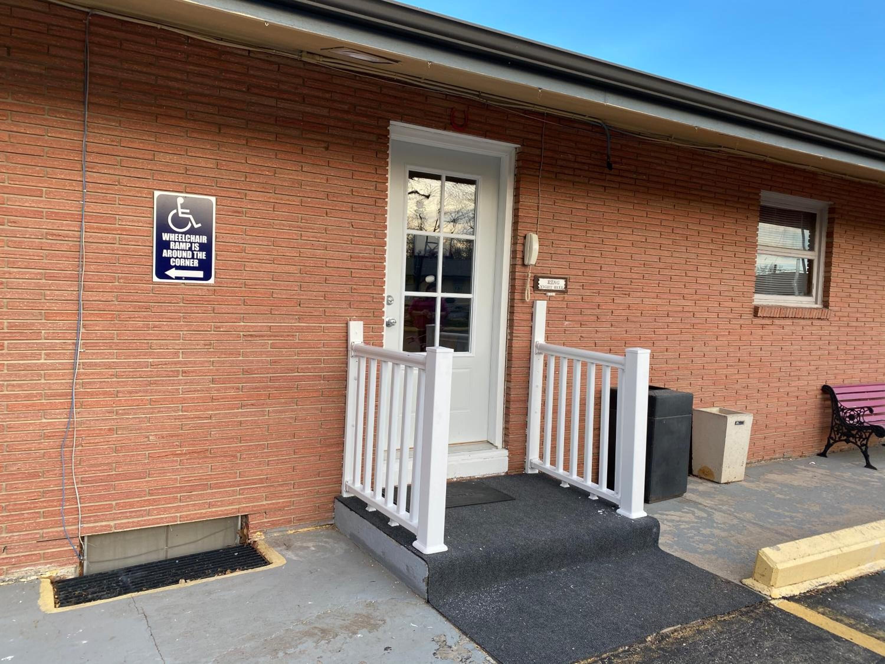 A brick building with a wheelchair ramp at the entrance, a white door, and a bench outside.