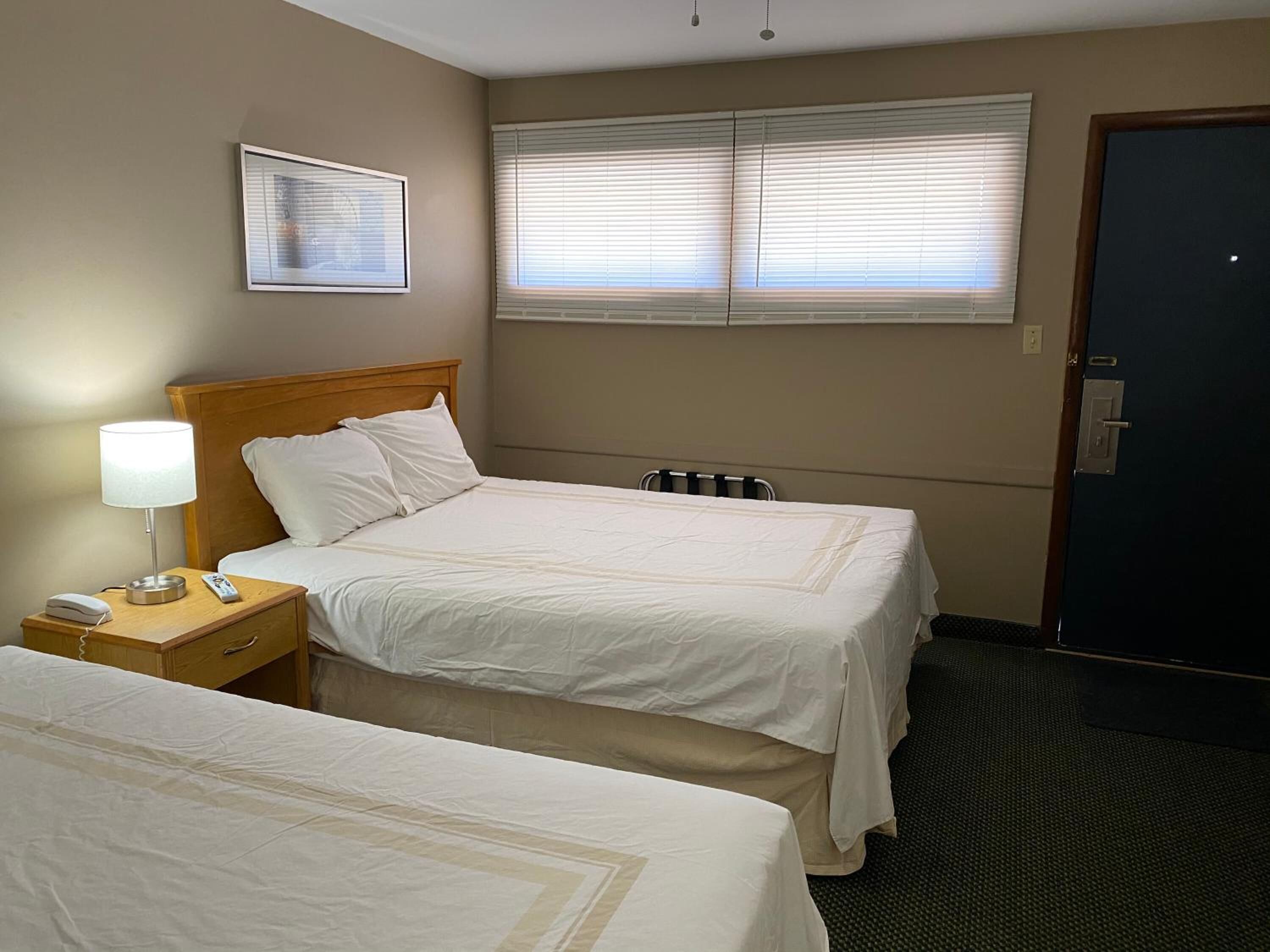A hotel room with two beds, a nightstand with a lamp, a framed picture on the wall, and a window with blinds.