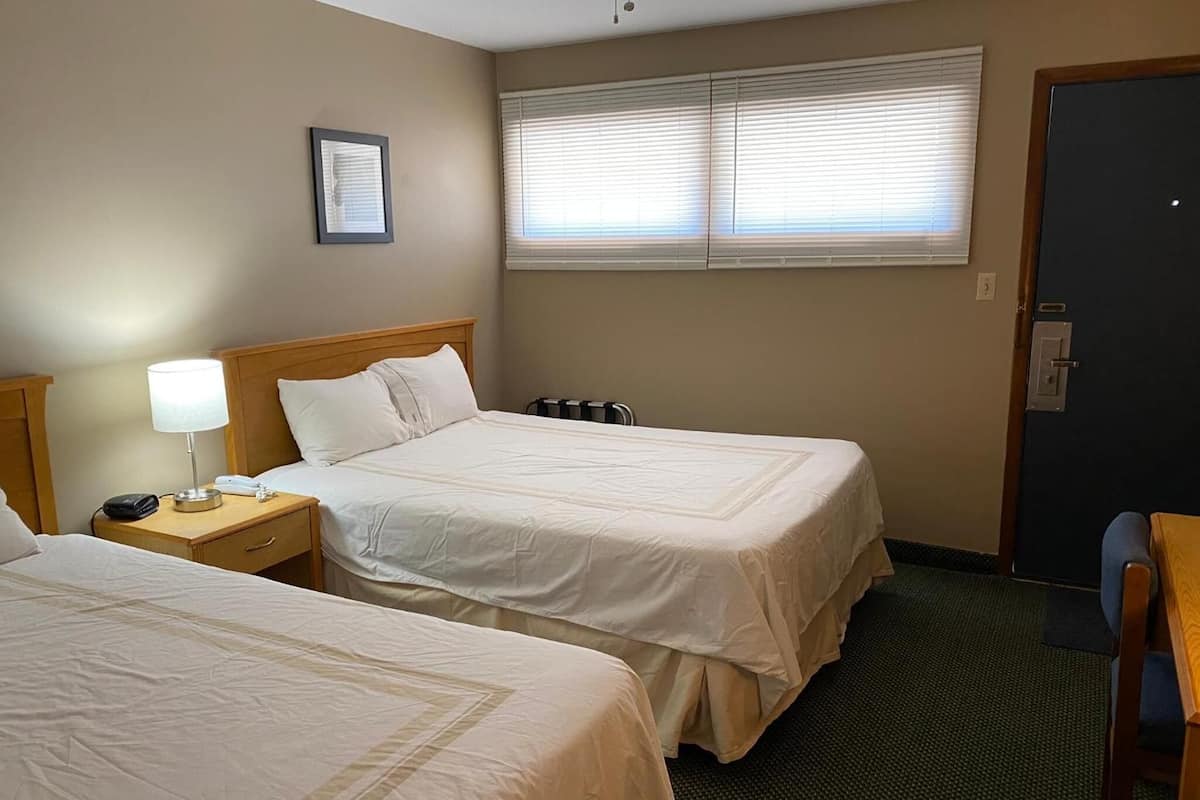 Ein Hotelzimmer mit zwei Betten, einem Nachttisch mit Lampe, einem gerahmten Bild an der Wand und einem Fenster mit Jalousien.