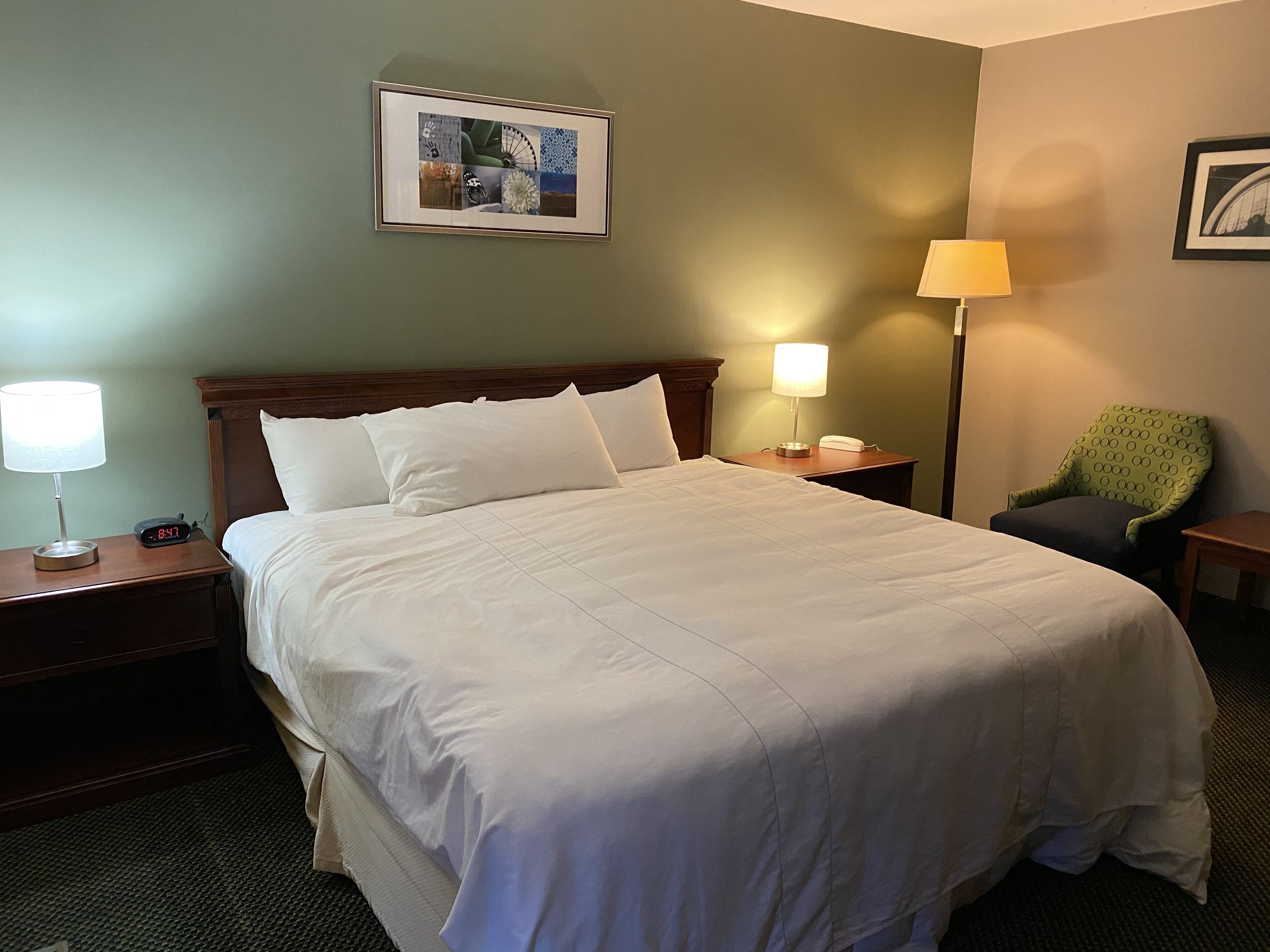 A neatly made bed with white linens, a bedside lamp, a clock, and a framed picture on the wall.
