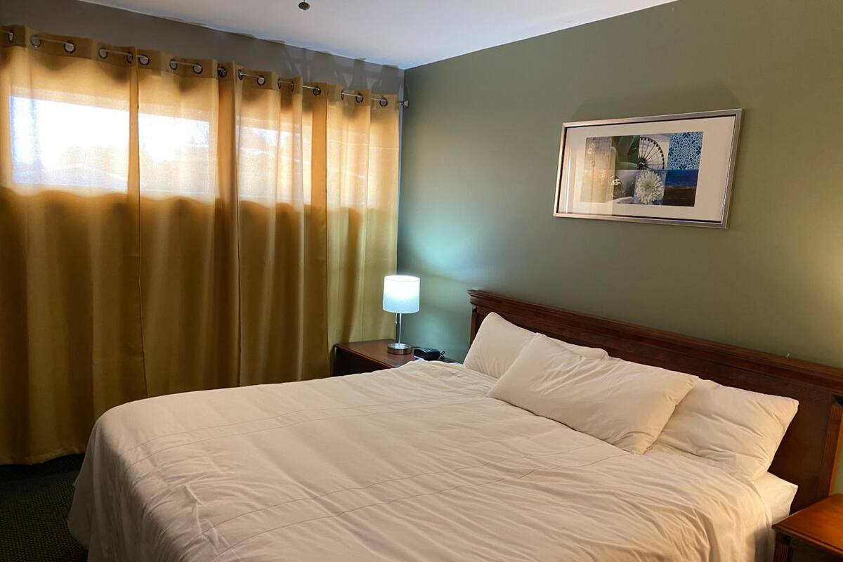 Ein Hotelzimmer mit einem großen Bett, einem Nachttisch mit Lampe, einem gerahmten Bild an der Wand und einem Fenster mit Vorhängen.