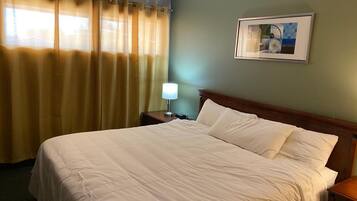 Una habitación de hotel con una cama grande, una mesita de noche con lámpara, un cuadro enmarcado en la pared y una ventana con cortinas.